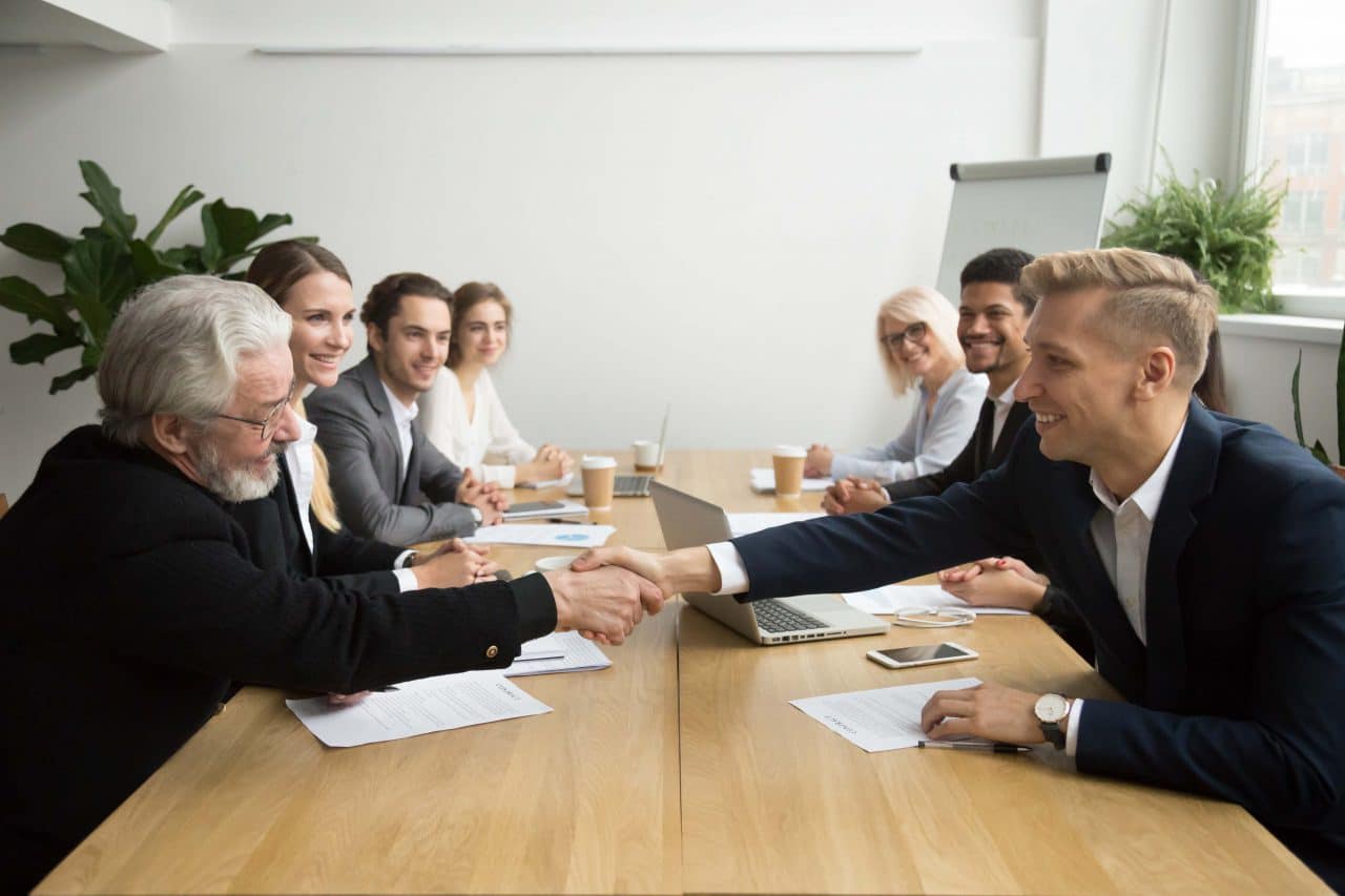 Young and old male partners shaking hands after signing contracts at group multiracial meeting, senior investor buying startup promising support handshaking entrepreneur, partnership deal concept